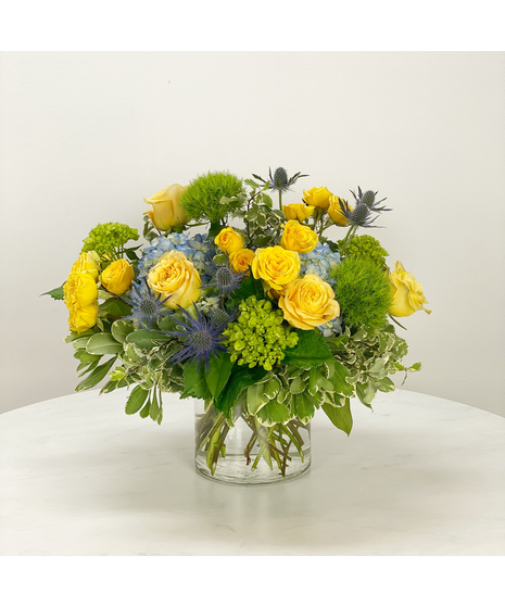 Bowl of blue and green hydrangea dotted with yellow standard and spray roses.