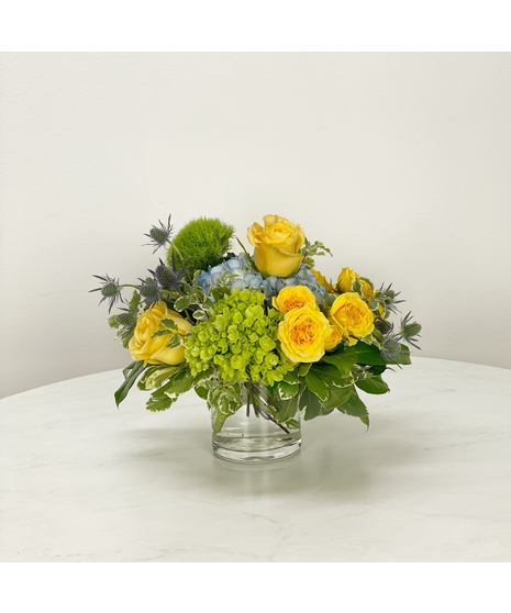 Bowl of blue and green hydrangea dotted with yellow standard and spray roses.