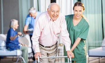 An elderly man using a walker is assisted by a smiling woman in green scrubs, while other seniors are seen in the background engaging in activities at a care facility.
